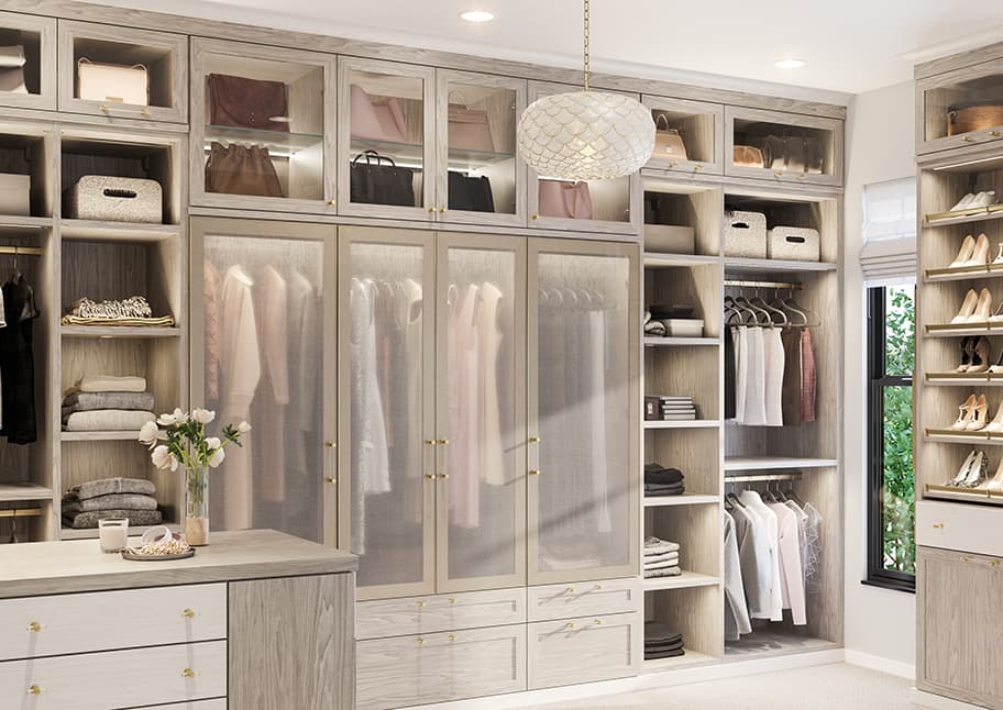 Walk in closet designed with glass cabinet doors, vanity, island and shoe storage by California Closets